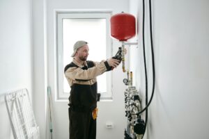 A person adjusting plumbing equipment near a window, wearing a cap and work uniform, demonstrates an efficient heating solution that aligns with an ympäristöystävällinen approach.