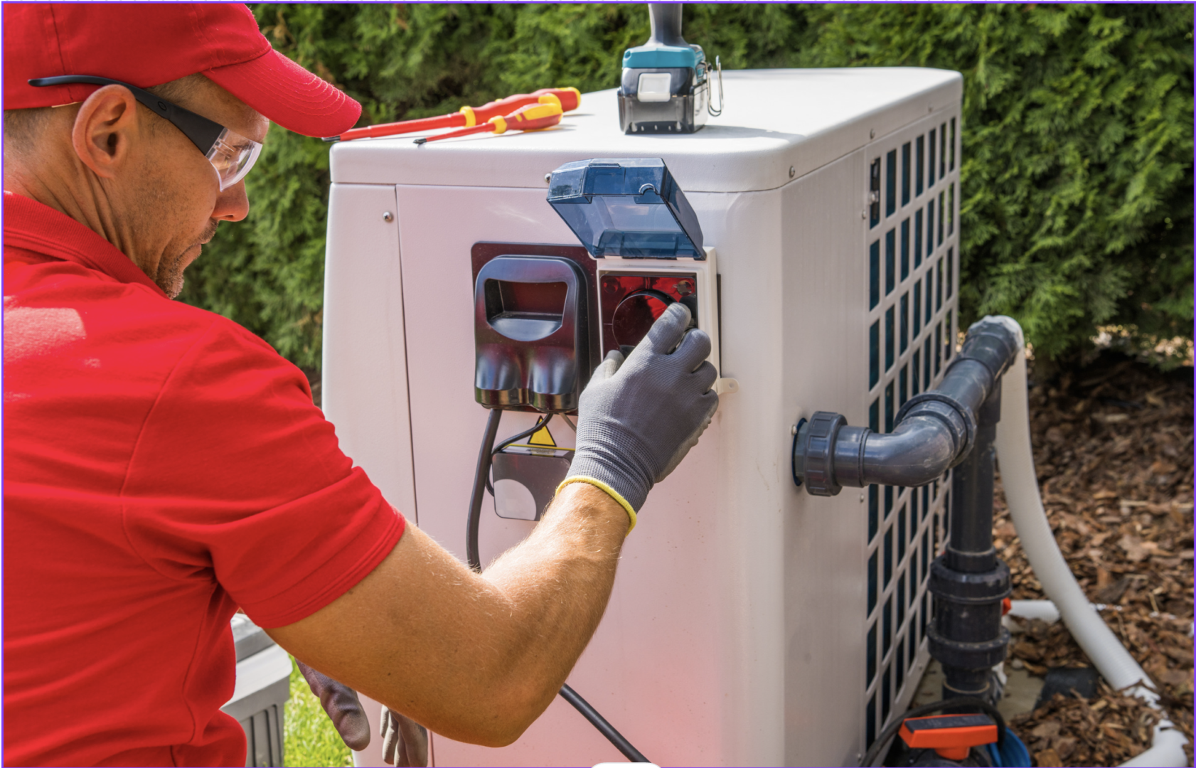 A technician in a red shirt and cap works on an outdoor ilmavesilämpöpumpun, adjusting the controls with tools visible on top of the unit during the asennus.