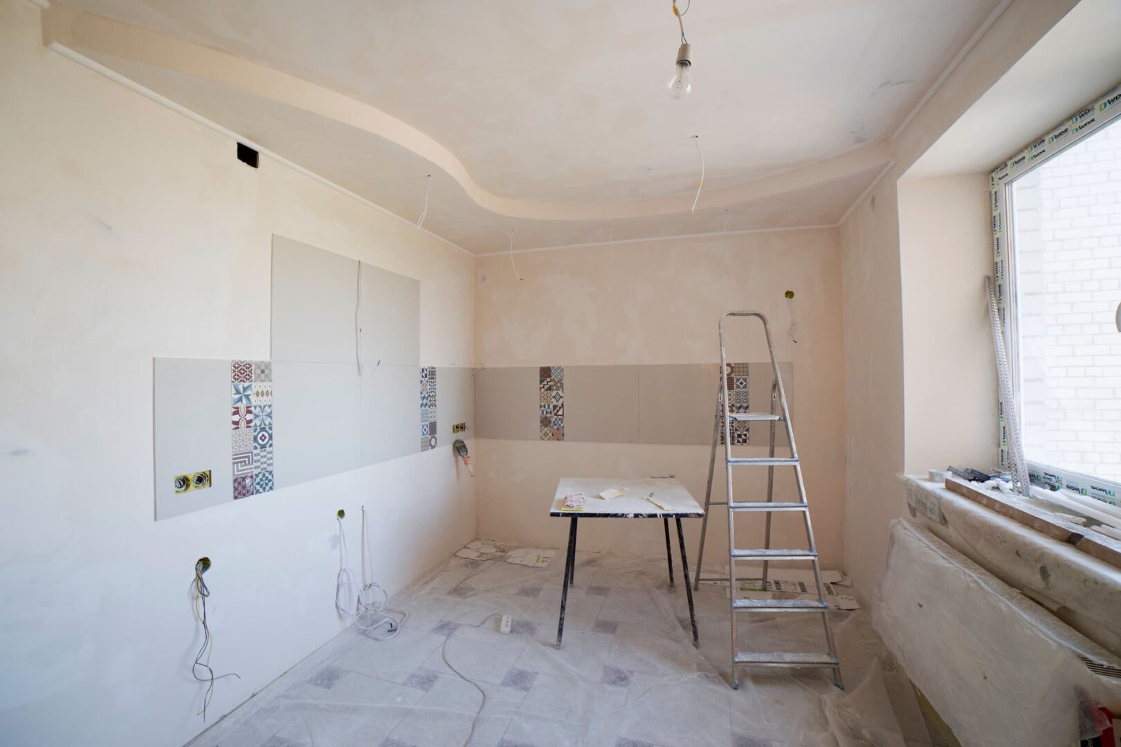 Kitchen renovation in progress: tile panels being installed, exposed wiring, and a metal ladder in a bright room